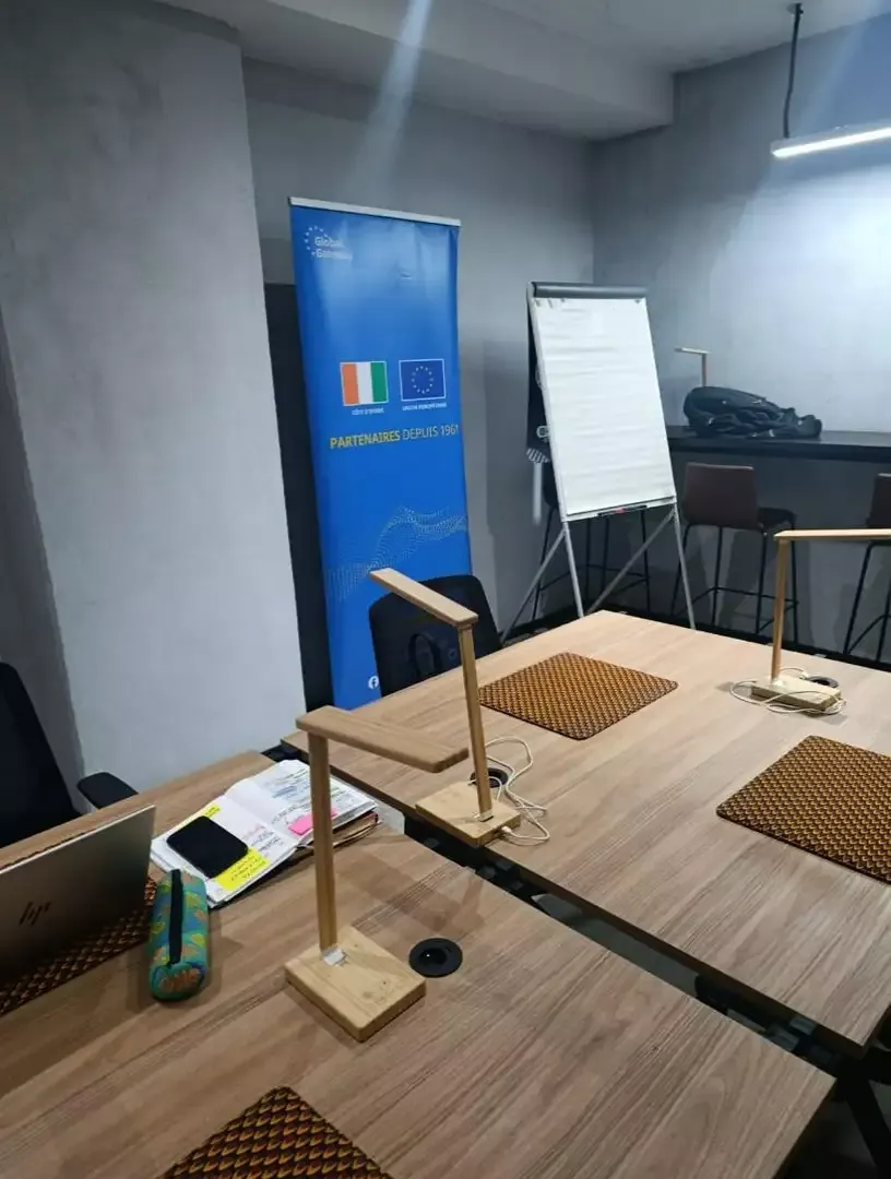 Vabicui — Une salle de réunion équipée d’une table en bois clair avec deux tapis ergonomiques, un support d’éclairage LED artisanal, un tableau blanc