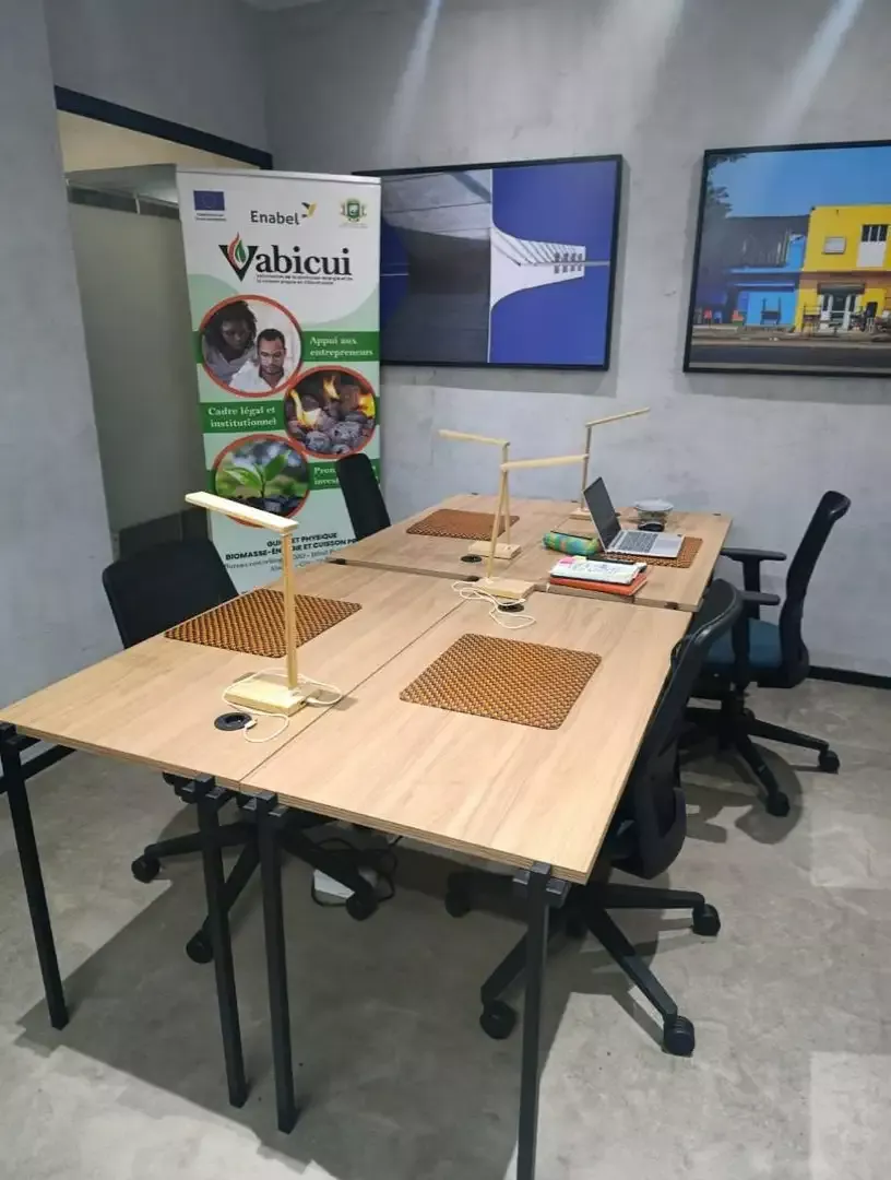 Vabicui — Un espace de travail moderne avec une grande table en bois, deux lampes solaires posées dessus, des chaises ergonomiques et un panneau promo
